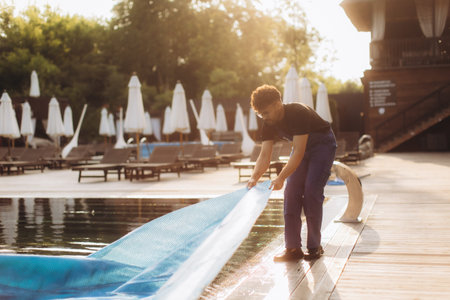 Pool maintenance worker removing a solar cover from a hotel pool in the morning sunshine, preparing for a refreshing summer dayの写真素材