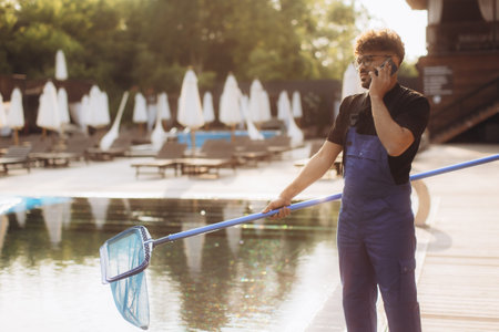 Pool cleaner wearing blue uniform, holding a landing net and talking on the phone next to a swimming...