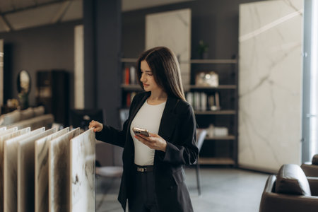 A confident woman in business attire examines marble samples while holding a mobile phone in a contemporary showroom.の写真素材