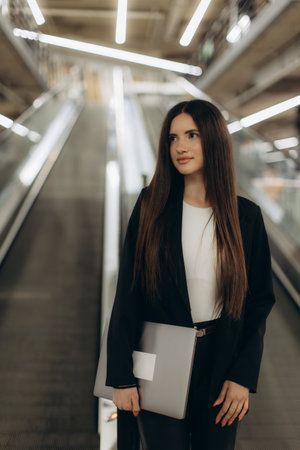 Professional young woman in formal attire holding a laptop, exuding confidence and focus while standing near an escalator in a sleek contemporary building setting.の写真素材