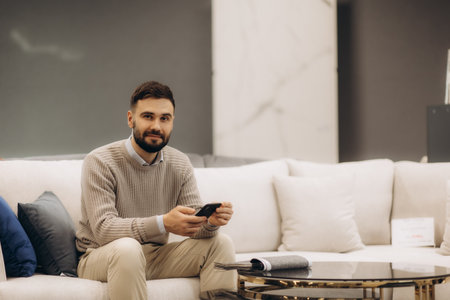 A man wearing a sweater, sitting on a couch, smiling warmly while holding a smartphone.の写真素材