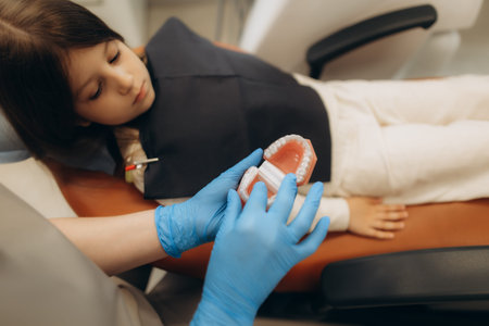 Dentist wearing blue gloves showing a teeth model with braces to a young patient lying on the...