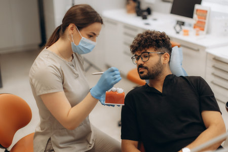 Female dentist explaining dental treatment to male patient using a teeth model in modern dental...