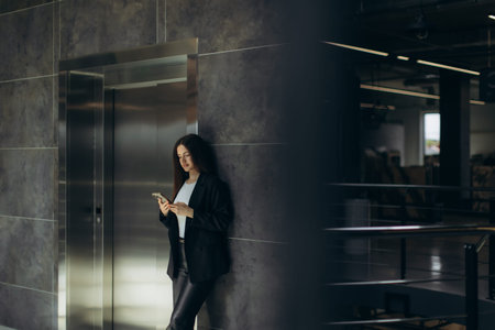 A business professional standing near an elevator in a contemporary office, using a smartphone. The...