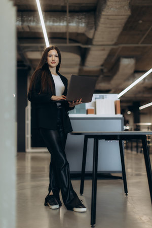 A professional woman standing in a contemporary office, holding a laptop and smiling confidently....