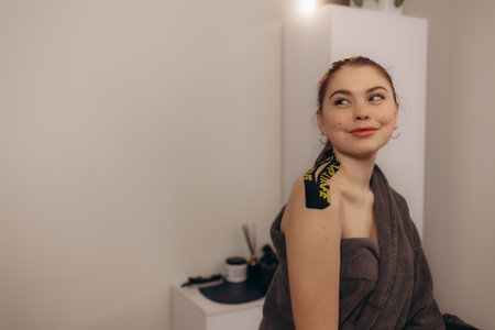 A young woman is smiling while wearing kinesio tape on her shoulder, seated in a cozy indoor space. The image exudes warmth, comfort, and well-being.の写真素材