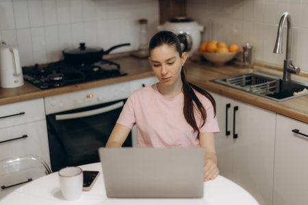 A young woman sits in a contemporary kitchen, focusing on her laptop. Coffee and a phone are nearby, enhancing the cozy and productive atmosphere.の写真素材