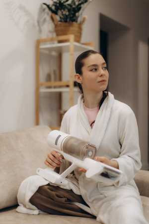 Young woman wearing a cozy white bathrobe using a wireless vacuum cleaner in a home setting. The background includes household furnishings such as shelves and decorative plants, emphasizing a neat and tidy environment.の写真素材