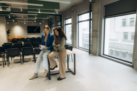 Two women engaged in a professional discussion holding documents while standing in a modern office...