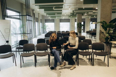 Two women engage in a conversation, sitting on chairs in a spacious conference room with plants and contemporary design, illustrating communication, professional interaction, and a modern work environment.の写真素材
