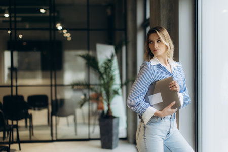 A stylish woman stands holding her laptop in a contemporary office environment, showcasing professionalism and modern work culture.の写真素材