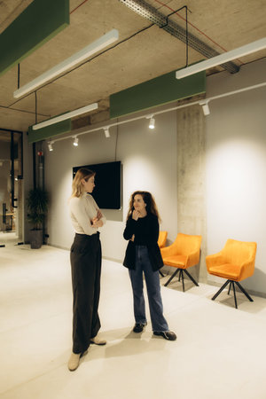 Two women stand and engage in a conversation within a well-lit, modern office, fostering ideas.