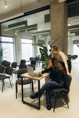 Two women focusing on a laptop in a contemporary workspace with chairs and a plant, emphasizing...