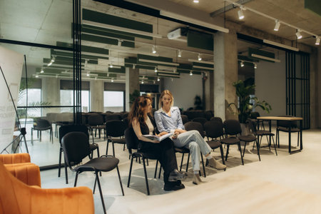 Two women sit in a contemporary conference room, engaging in discussion, with chairs and glass...