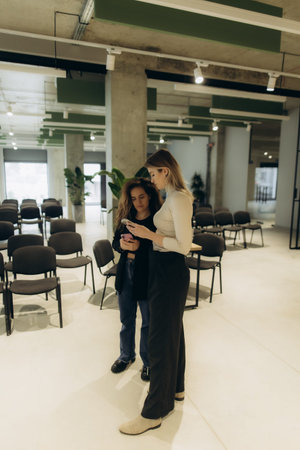 Two women are seen in conversation in a modern office setting featuring minimalist decor and ample...