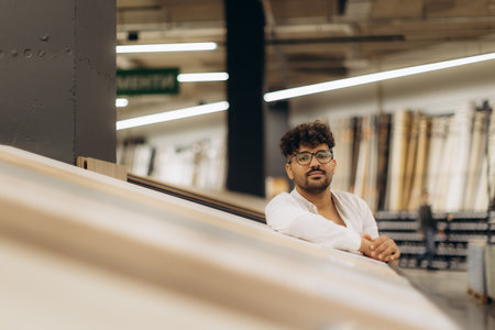 A young man wearing glasses leans on a counter in a well-lit, contemporary store, reflecting confidence and style. Modern fixtures provide ambiance, creating an inviting atmosphere for shopping and design exploration.の写真素材