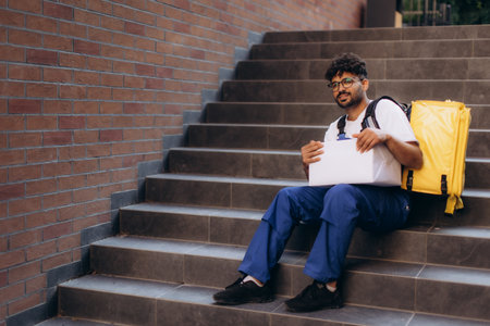 Delivery man resting on stairs, sitting with a package in hand, wearing a yellow backpack, enjoying a brief moment of respiteの写真素材