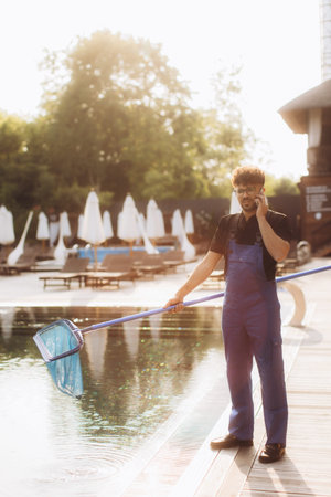 Pool cleaner wearing blue overalls holding cleaning net and talking on the phone by the poolside on...