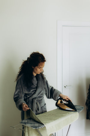A person in comfortable attire ironing clothes in a home setting. The depiction conveys a sense of daily chores and homely atmosphere, highlighting simplicity and routine.の写真素材