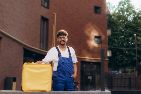 Delivery man holding a bright yellow thermal backpack while standing on a bustling city street next to a modern buildingの写真素材