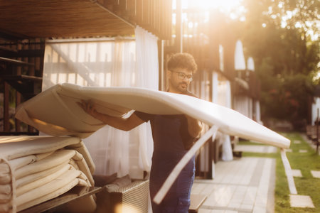 Hotel employee carrying a white mattress outdoors, positioned near flowing white curtains as the sun sets, casting warm light across the sceneの写真素材