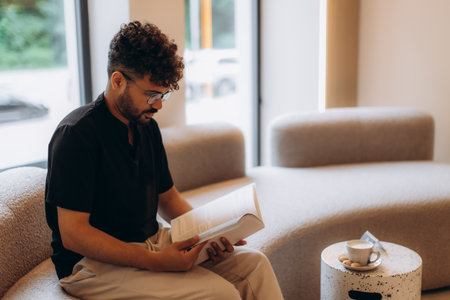 Young businessman is sitting on sofa in office lounge, reading document and enjoying cup of coffee during breakの写真素材