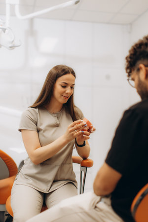 Female dentist explaining dental treatment to male patient using a teeth model in a modern dental...
