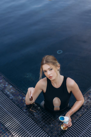 Young woman in black swimsuit relaxing in a luxury pool with blue cocktail and sunglasses, enjoying a moment of leisureの写真素材