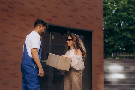 Delivery man handing over a cardboard box to a smiling woman, both enjoying a moment of satisfaction in front of a charming brick buildingの写真素材