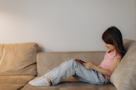 A young girl sitting comfortably on a sofa, using a tablet device, showcasing focus and leisure.の写真素材
