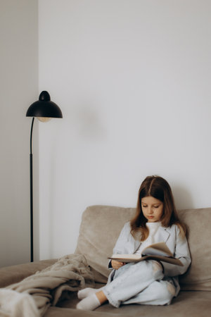 A young girl is seated on a cozy couch with a book in her hands, reading attentively in a serene and tidy indoor setting.の写真素材