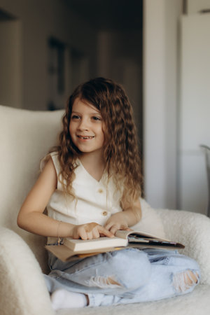 A smiling young girl is seated indoors on a cozy chair while reading a book.の写真素材