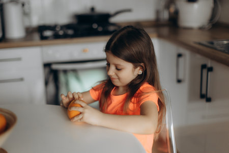 A young girl is seen peeling an orange while sitting at a table in a kitchen. Natural light creates a warm and comfortable atmosphere, emphasizing the simplicity and joy of everyday activities.の写真素材