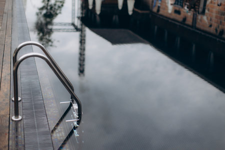 Metal grab bars reflecting in the still water of an empty rooftop swimming pool, creating a serene...