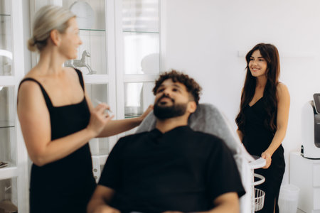 Young man receiving a cosmetic procedure from a female cosmetologist while another female assistant...