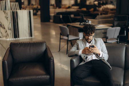 A businessman sits in a contemporary office environment, engaged with his smartphone. The setting features modern decor, leather chairs, and other office equipment, portraying a casual yet professional setting.の写真素材