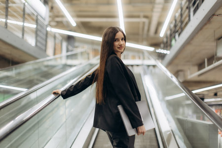 Confident businesswoman in formal attire holding a laptop while riding an escalator in a contemporary office environment. The image conveys professionalism, elegance, and a sense of corporate professionalism.の写真素材