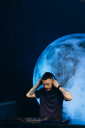 A DJ wearing headphones performing live at night with a large illuminated moon behind. Capturing an energetic, vibrant, and inspiring artistic moment during an exceptional and visually compelling music event.の写真素材