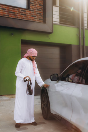 Man in traditional thobe and keffiyeh preparing to charge his modern electric car charger at his houseの写真素材