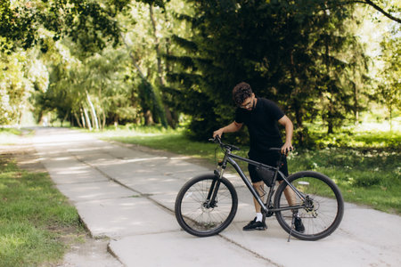 Young man pausing on a concrete path in a lush green park, holding his bike. Enjoying summer day outdoorsの写真素材