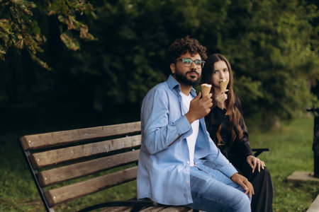 Diverse couple sharing moment, eating ice cream cones on park bench. Enjoying leisure outdoors, relaxation, and sweet refreshmentの写真素材