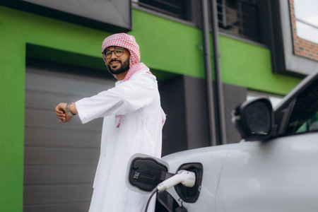 Arab man in thobe and keffiyeh waiting, checking his watch while electric vehicle is charging at a green stationの写真素材