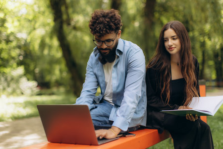 Young diverse students using a laptop and a notebook while studying together on a bench in a park, focusing on educationの写真素材