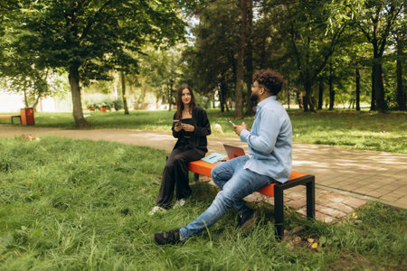 Young students discussing and using smartphones, studying outdoors on a park bench, enjoying break timeの写真素材