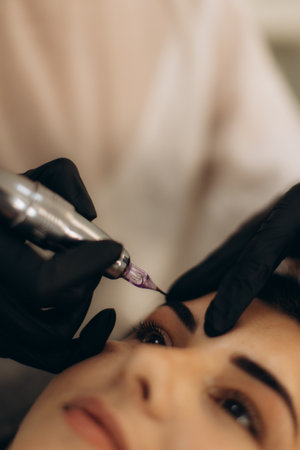 Specialist applying permanent makeup to a woman's eyebrows using a microblading machine, enhancing beautyの写真素材