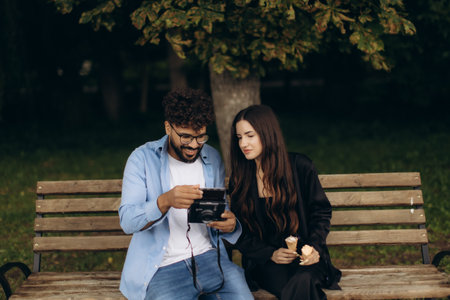 Young couple sitting on a park bench, looking at an instant photo together and enjoying ice cream during a relaxed dateの写真素材