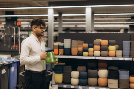 A man in a store aisle exploring and carrying a green watering can, while observing stacked assorted colored plant pots, exemplifies home gardening or interior decoration shopping.の写真素材