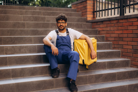 Courier resting on stairs after delivering a food order, taking a break with a yellow insulated thermal backpack beside himの写真素材
