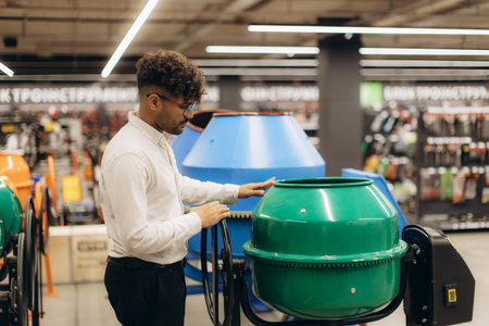 A man examines a green concrete mixer in the tool department of a hardware store. The store features a modern layout with various construction equipment and tools available for shopping.の写真素材