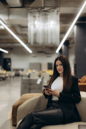 A confident woman sits on a stylish chair holding a smartphone, located in a modern, well-lit interior. She exudes professionalism and ease in the contemporary environment.の写真素材